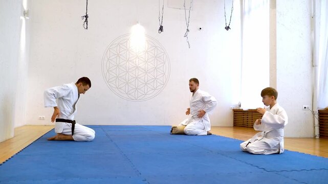 Three young martial artists in white gi sit cross-legged on a blue mat. They practice controlled movements with calm, focused expressions. Geometric design adorns the clean white wall behind them