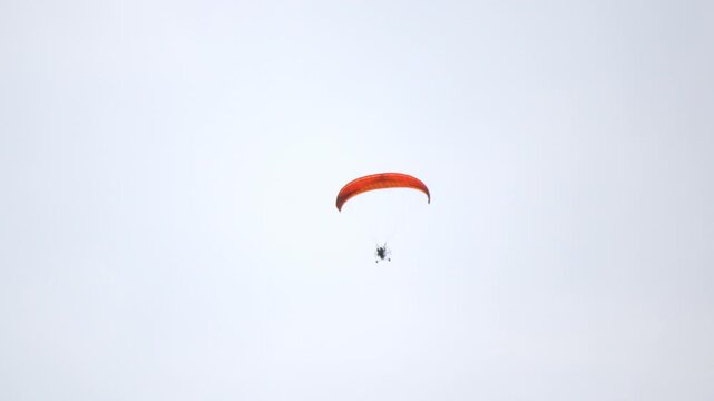 Parachute soars high against pale sky. Red canopy glides gracefully above. Small figure floats peacefully in air. No wind, no rush, no stress shown. Ideal for tranquil travel or skydiving themes
