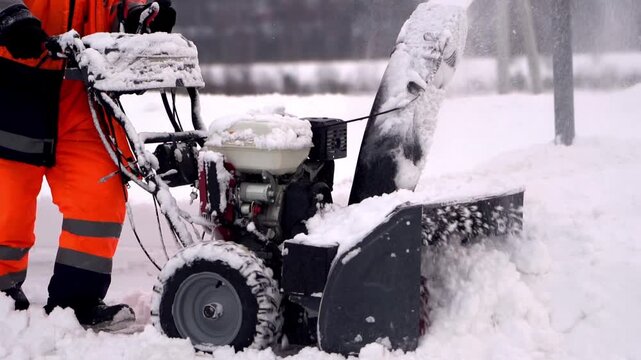Worker clears snow with mechanical snowblower. Snowflakes swirl in cold, wintry air. Person wears bright orange safety gear. Equipment sprays snow forward powerfully