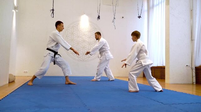 Three martial artists move together on a blue mat. One wears a black belt, the others wear white. They train inside a bright, minimalist studio. Disciplined focus and coordinated movement