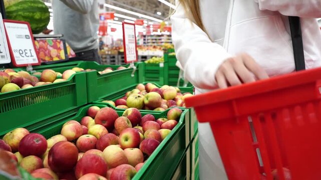 Shopper picks apples in a supermarket aisle. The woman's hand selects a fruit from a crate. Her basket sits nearby, ready to hold more goods. No emotions are visible, just routine grocery activity