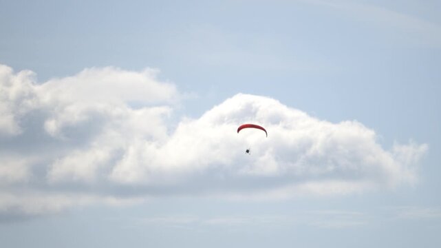 Parachute glides above soft white clouds. Sky is vast and calm with gentle blue tones. Red canopy contrasts serene background. Parachutist floats freely, unattached to ground