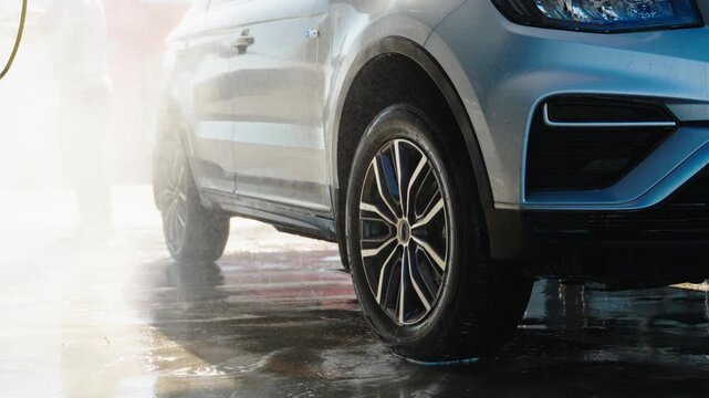 Water sprays clean a modern vehicle's lower body. Wet ground reflects the bright, dynamic cleaning spray. Chrome wheels gleam under the force of the jet