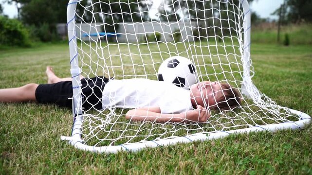 Child rests inside a soccer goal, smiling. Football lies beside them on grass. Lush green field surrounds the scene. Playful moment captured outdoors. Ideal for sports, childhood