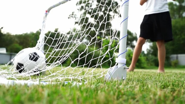Footage captures a child's joyful soccer goal moment. Ball nestled in net, green grass beneath. Child runs away, bare feet touching earth. Sunlight filters, trees line distant background