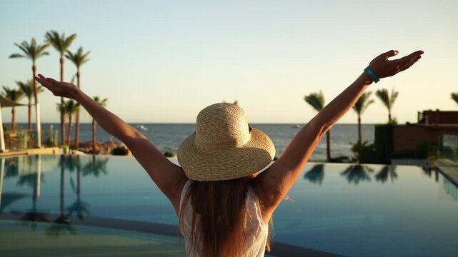 Sunset glow washes over calm pool and ocean. Woman stands back, arms raised, joyful, free. Tall palms frame horizon, reflecting in still water. Serene beachside escape. No animals or plants identified