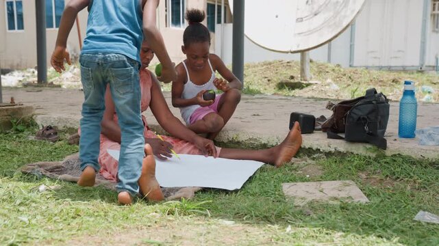 Woman draws outdoors surrounded by grass and scattered objects. Children engage in separate activities nearby creating casual family scene with art materials