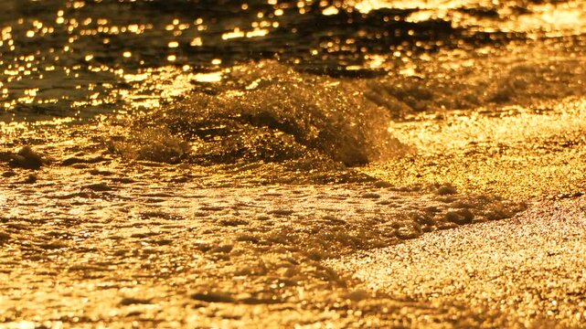 Golden sunset reflections dance across rolling sea surf as tide water cascades onto a coastal shore. Static high-angle close-up shot.