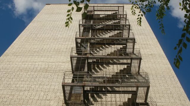 Low angle view of an old external metal fire escape staircase casting shadows on a tall white brick building facade against a blue sky with clouds, showing an urban architectural detail
