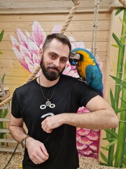 Vertical medium shot of a bearded Caucasian man smiling with a large blue and yellow macaw parrot sitting on his shoulder in a zoo aviary against a painted wooden wall background © Tina