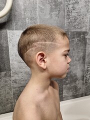 Vertical profile portrait of young boy with short blonde haircut featuring shaved lightning bolt design on side standing in bathroom against grey tiled wall under indoor light © Tina