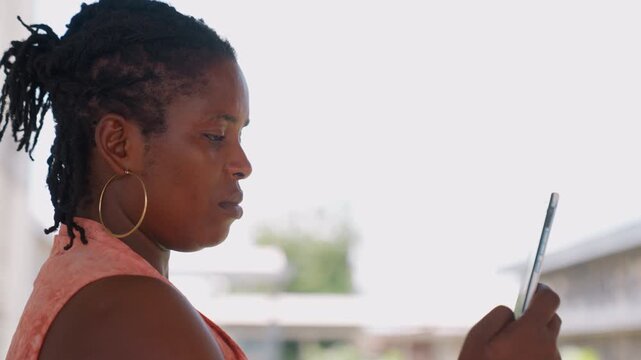 Woman with dreadlocks wearing large hoop earrings and orange top holds smartphone looking at screen in bright outdoor light. Her posture suggests engagement or focus on digital content
