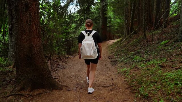 Woman with white backpack hiking along tranquil forest trail, walking deeper into lush green woodland on peaceful summer day outdoor nature adventure