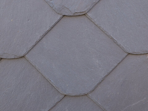 Grey slate shingles form a regular honeycomb pattern on a facade. The split surfaces and irregular edges reveal the natural stone's characteristic texture.