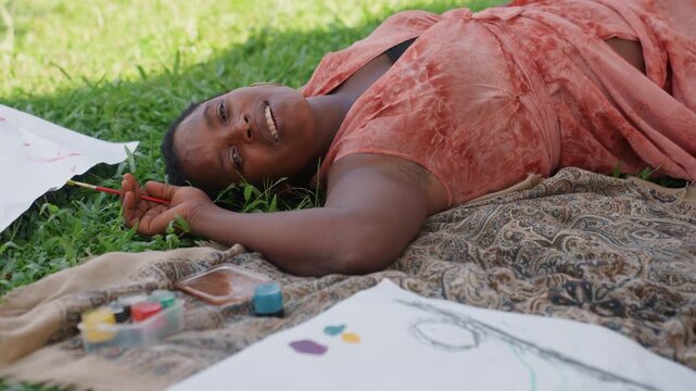 Woman lies outdoors holding paintbrush near paper with watercolor paints spread nearby. Woman rests comfortably as part of artistic activity in nature