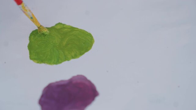 Close up view of green and purple paints being applied to white surface. brush spreads colors creating visible texture details. scene focuses on paint application