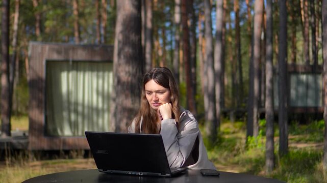 Cinematic slider tracking shot from right to left of a young woman working on a laptop at a forest eco resort terrace.