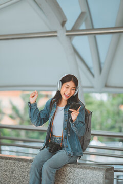 Happy young Asian woman traveler listening to music with headphones while using smartphone and raising fist in excitement, sitting with backpack and camera.