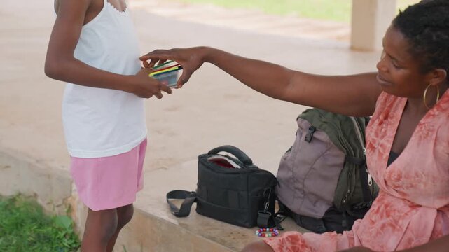 Woman hands art supplies to young girl outdoors on concrete steps. interaction involves handing over colored pencils with bags and beaded bracelets visible in setting