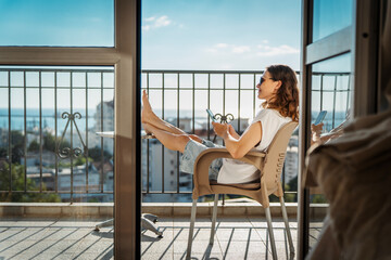Fototapeta premium Happy young woman sitting on balcony chair with barefoot legs on table and looking at smartphone screen.