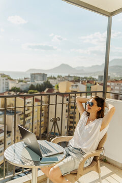 Relaxed young woman sitting on balcony with laptop and hands behind head enjoying city view.