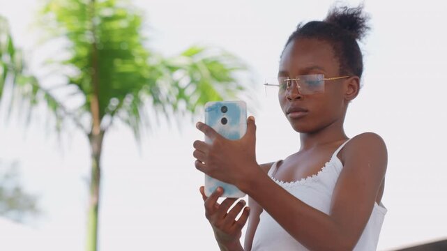 Shows child interacting with mobile device outdoors. scene includes subject hands gripping electronic gadget against bright natural light suggesting casual use in daylight