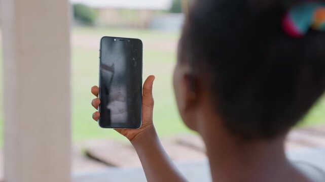 Young girl holds up her smartphone indoors while facing away from camera. background shows blurred outdoor scene visible beyond window suggesting daytime with natural light filtering