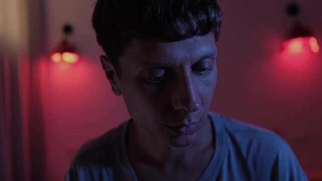 Anxious man overwhelmed by an unexplained sense of danger, looking down and reacting with fear as if expecting something bad to happen in a dark bedroom with red and blue light