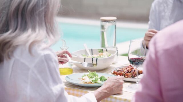 Group of elderly friends gathered around a table for a cheerful outdoor lunch. A woman with grey hair eats salad and drinks red wine