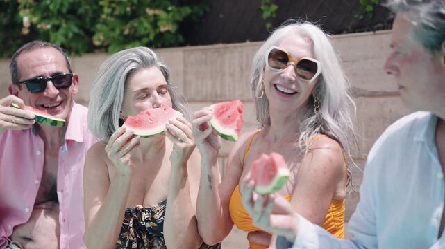 Happy senior friends laughing and eating fresh watermelon slices. Cheerful mature people enjoying a sunny summer day together at the poolside