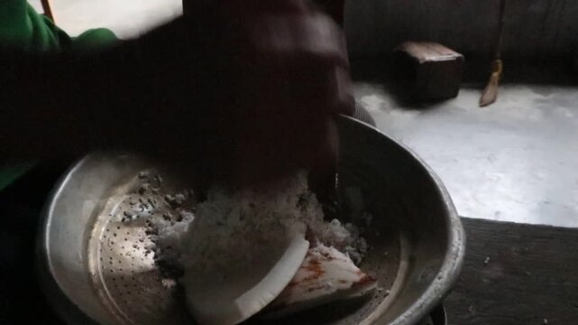 Traditional hand grating fresh coconut with a manual coconut grater for food preparation in a rural Indonesian kitchen.