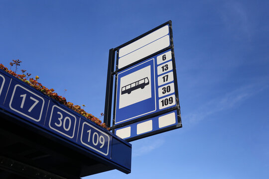 Blue public bus stop sign under clear sky. Route numbers on urban transport shelter. Passenger transit infrastructure background. Municipal bus station marker. City travel symbol. Street detail.