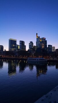 Vertical view of the illuminated skyline with its skyscrapers at blue hour, city lights reflecting in the river Main, seen from the Eiserner Steg footbridge, Frankfurt am Main, Hesse, Germany