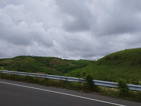 Scenic roadside view of rolling green hills and grassy slopes behind a metal guardrail under overcast cloudy sky.
