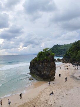 High angle view of a huge rock formation on a sandy beach with tourists and rolling ocean waves under overcast sky.