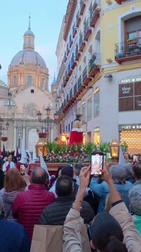 Vertical view of a Holy Week procession float with a statue of Christ, flowers and lanterns passing a crowd filming with phones at dusk near the Basilica del Pilar, Zaragoza, Aragon, Spain