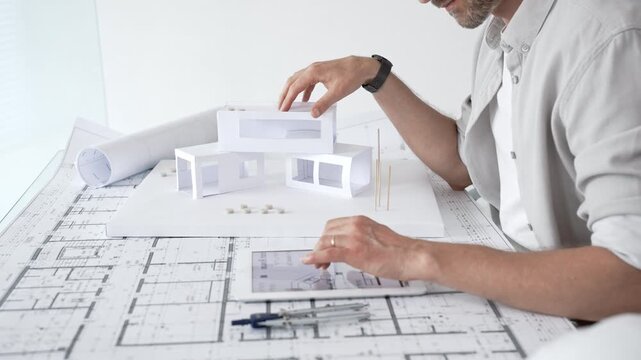 Architect working on a new building project, using a digital tablet for design and planning, surrounded by blueprints and a conceptual scale model of a modern house