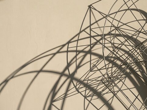 Abstract composition of a geometric metal wire sculpture casting complex, elongated shadows onto a smooth, light beige plastered wall surface with high contrast natural lighting.