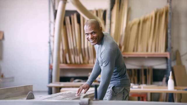 Male woodworker carefully positions a plank on a table saw, then turns to smile confidently at the camera in his carpentry workshop