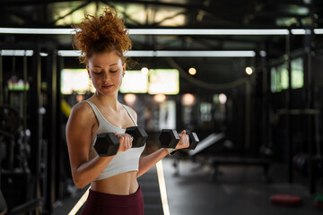 Naklejka na ściany i meble Young woman lifting dumbbells performing bicep curls in gym