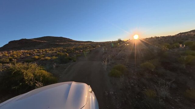 A white car is driving along a narrow, bumpy road with a poor surface in the African savanna. Sunset time.