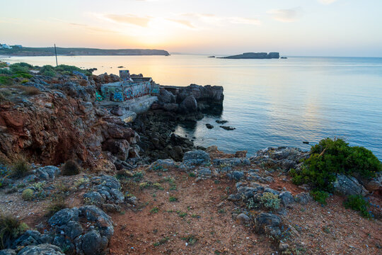 A golden sunset glows over calm ocean waters beside a rugged rocky coastline with red cliffs and low coastal shrubs. A weathered concrete ruin sits on the shore as distant headlands fade in soft haze