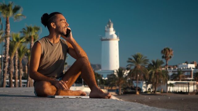 Smiling young man talking on a smartphone with a laptop on the ground while sitting cross-legged on a concrete seaside ledge by a white lighthouse, palm trees, remote work lifestyle