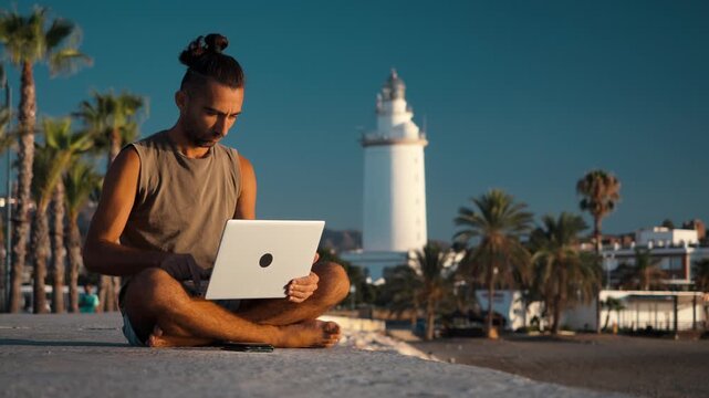 Young digital nomad sitting cross-legged on a concrete seaside promenade typing on a laptop with a white lighthouse and palm trees behind him, young man, remote working, laptop lifestyle
