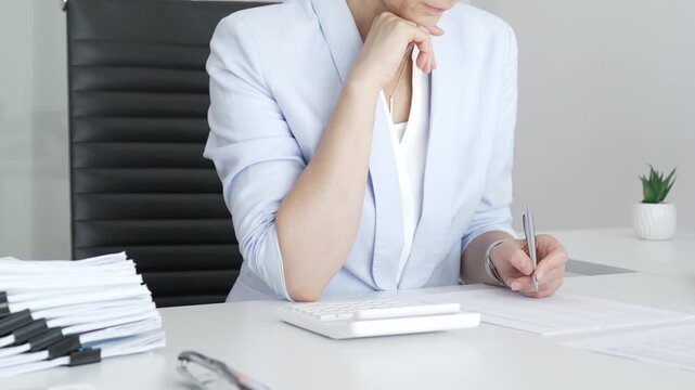 Professional woman working on financial documents, calculating data with a calculator and writing notes in a modern office, managing tax forms and business reports