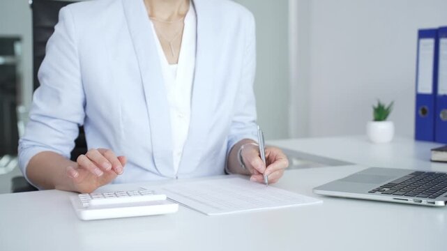 Professional woman working on financial documents, calculating data with a calculator and writing notes in a modern office, managing tax forms and business reports
