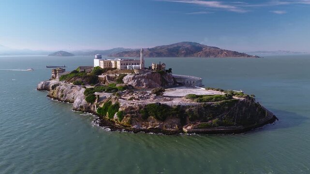 Aerial view of Alcatraz Island situated in San Francisco Bay, showing the former federal penitentiary, lighthouse, water tower, rocky cliffs, and surrounding Pacific waters under a clear sky.