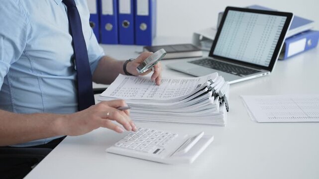 Businessman reviewing and organizing a large stack of financial reports, invoices and ledgers on a white desk with laptop spreadsheets, calculator and magnifying glass close up