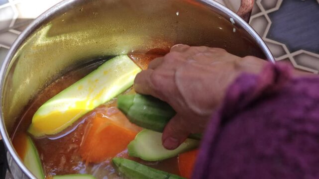Hand adding zucchini to boiling couscous broth with pumpkin in pot