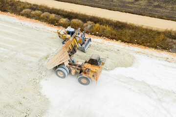 An aerial view of heavy machinery including a dump truck unloading soil, a bulldozer, and a grader working on a road construction embankment. © scharfsinn86